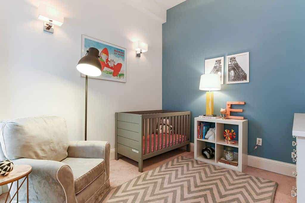This nursery is furnished with a wooden crib and taupe armchair lighted by a black dome floor lamp. It includes a chevron rug and open shelving topped with a yellow table lamp.