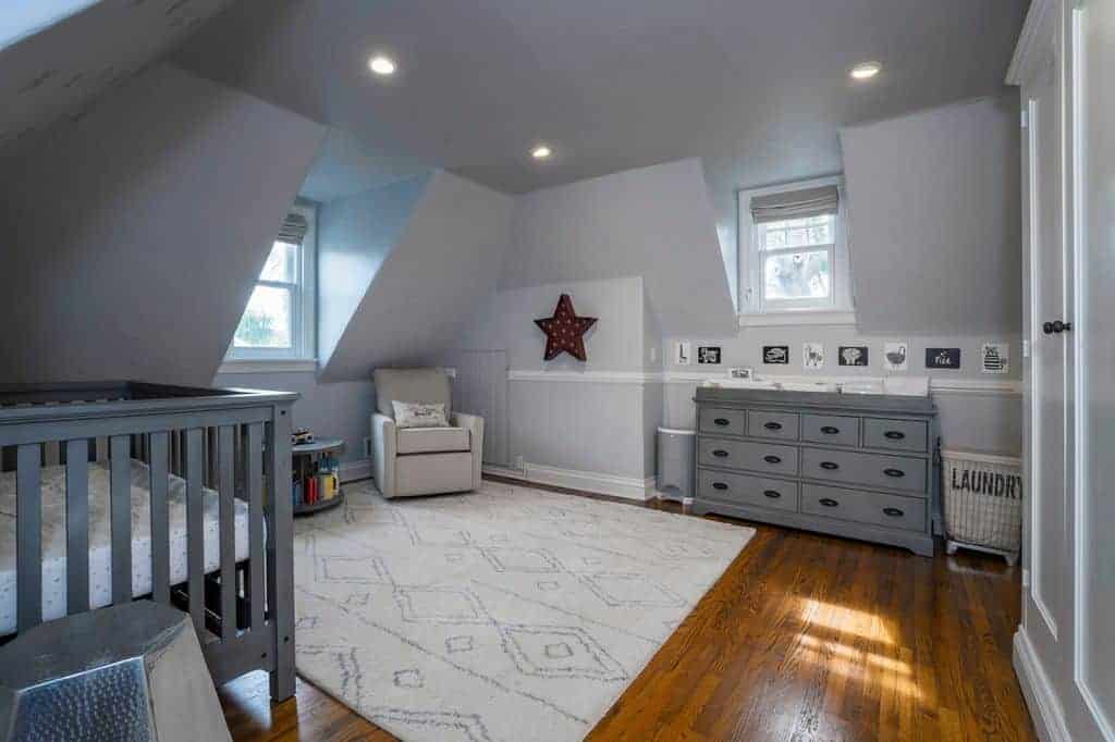 Gray nursery with vaulted walls and carpet flooring topped by a patterned area rug. It includes an upholstered armchair and crib that matches the dresser flanked by trash and laundry bins.