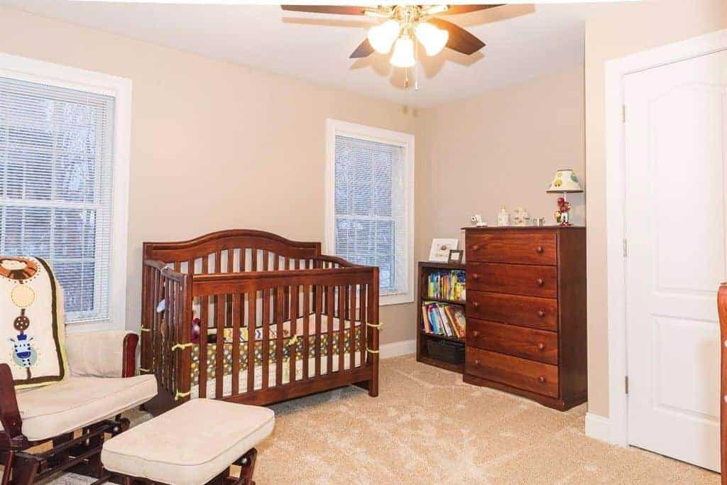 A wooden crib complements the drawer chest and cushioned chair paired with a matching ottoman in this nursery with carpet flooring and ceiling fan with lights illuminating the entire space.