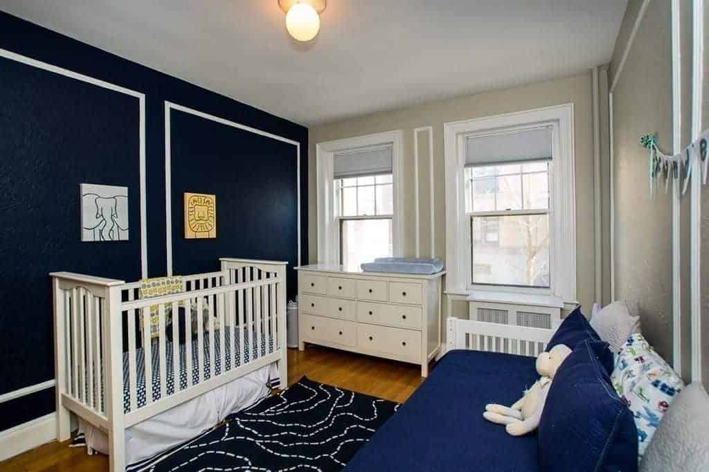 A skirted crib matches the white seat topped with navy blue cushion and fluffy pillows. It includes a drawer chest and black printed rug that lays on the rich hardwood flooring.