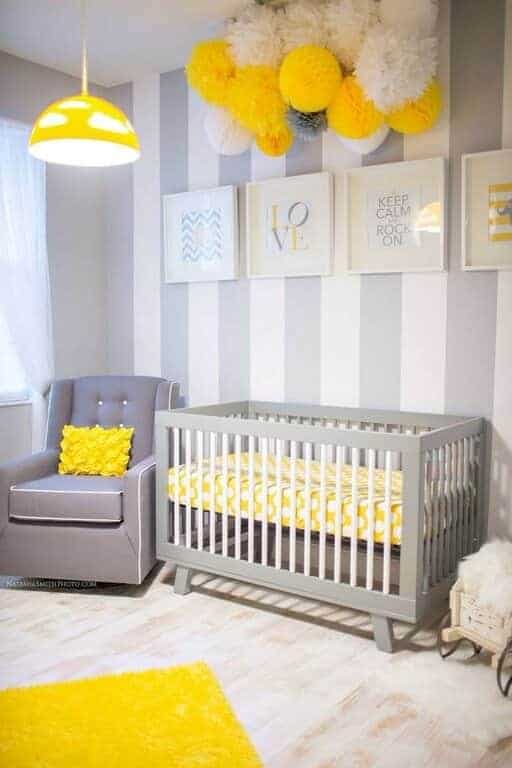 A yellow dome pendant light illuminates the gray armchair in this cheerful nursery decorated with pompoms and white framed artworks mounted on the striped accent wall.