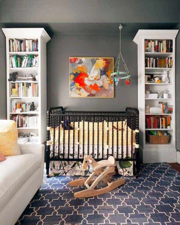 Colorful artwork hangs above the black crib flanked by white shelving units against the gray inset wall. It is accompanied by a cozy sofa and wooden rocking horse over the deep blue patterned rug.