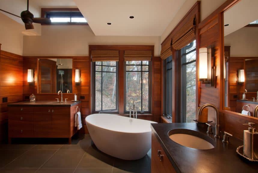 The white freestanding bathtub stand out against the rest of the bathroom that is dominated by the redwood elements on the window frames and walls that blends with the two wooden vanities illuminated by modern wall-mounted lamps.
