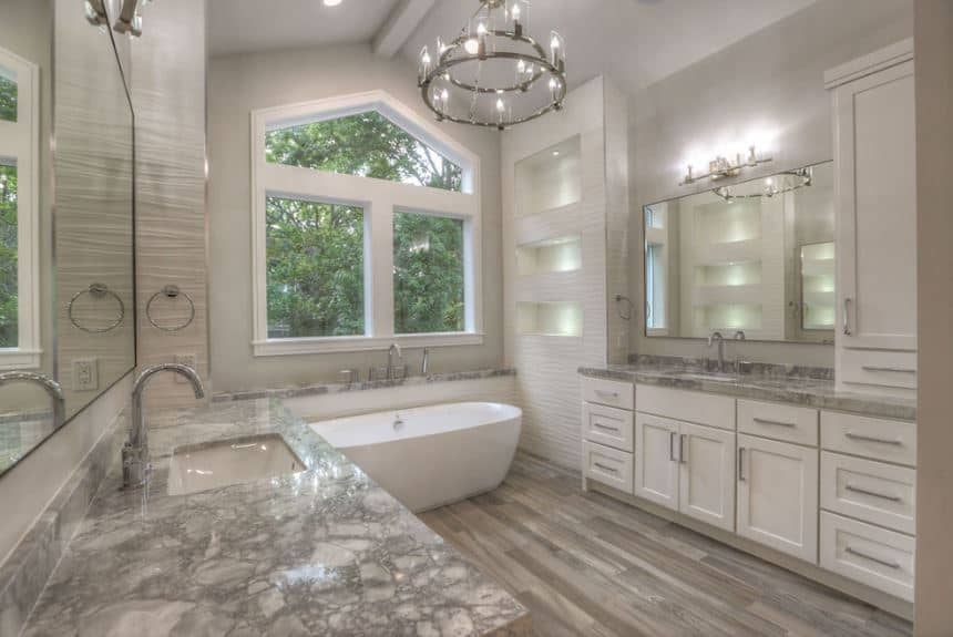The light gray cathedral ceiling has a single exposed beam running in the middle that supports the brilliant two-tier farmhouse-style chandelier matching the wall lamp above the mirror of the vanity that has white shaker cabinets and drawers.