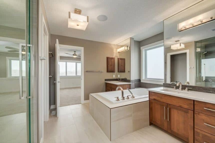 The white flooring tiles are contrasted by the two wooden vanities with white countertops and large wall-mounted mirrors that has modern lamps. These two vanities are flanking a white bathtub inlaid with the same tiles as the flooring.