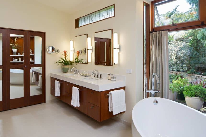 This craftsman-style bathroom features mirrored wooden folding closet doors adjacent to the wooden floating vanity that stands out against the white walls warmed by the yellow lights of the modern wall lamps beside the mirrors.