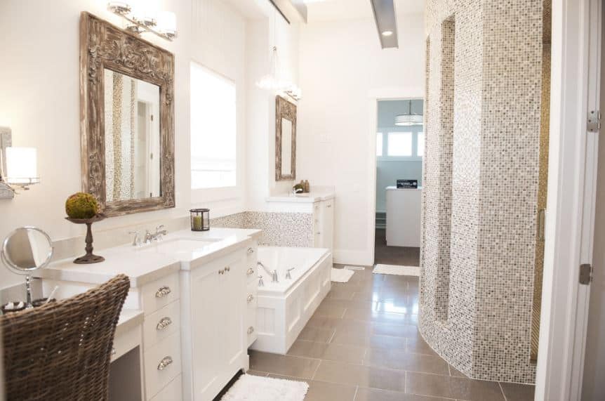 The gray flooring tiles of this bright bathroom is complemented by the complex patterned finish of the shower room and bathtub backsplash. The white walls and white vanities flanking the bathtub are complemented by the elegant wood frames of the mirrors.