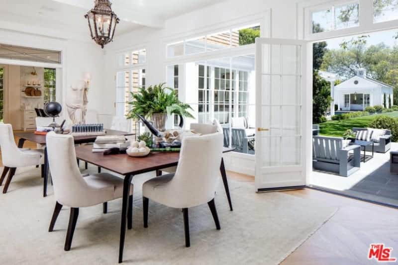 The patio scene outside the French glass windows and glass doors bring in natural lighting that brightens up the white ceiling with exposed beams that support a classic lantern-like pendant light that that brings a certain Spanish-style flair to the bright aesthetic.