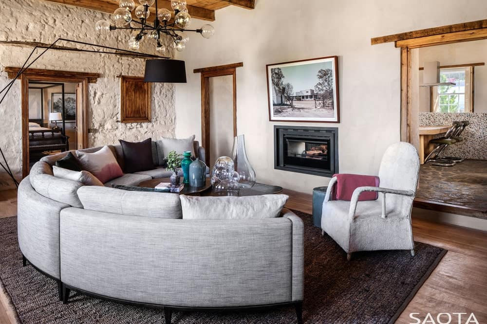 This is a simple living room with a large gray curved sectional sofa paired with a dark coffee table across from the fireplace. These are complemented by the chandelier and the beamed ceiling.