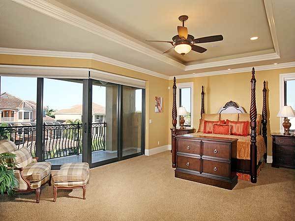 The spacious and bright primary bedroom has a alrge dark wooden four-poster bed that stands out against the beige walls, tray ceiling and beige carpeted flooring.