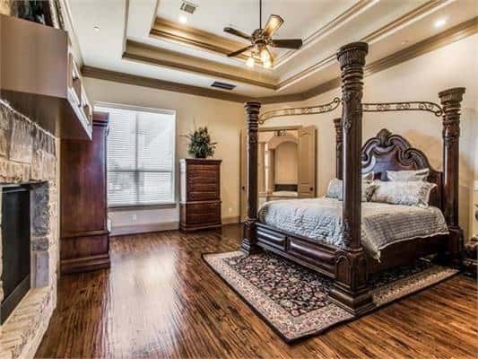 This is the primary bedroom that has a large dark wooden four-poster bed that matches the hardwood flooring and the large dresser by the stone fireplace.