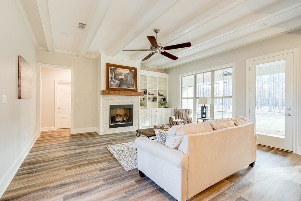 The beige sofa set of the living room matches well with the beige walls and beamed ceiling that has a ceiling fan over the coffee table across from the fireplace that is topped with a wall artwork.