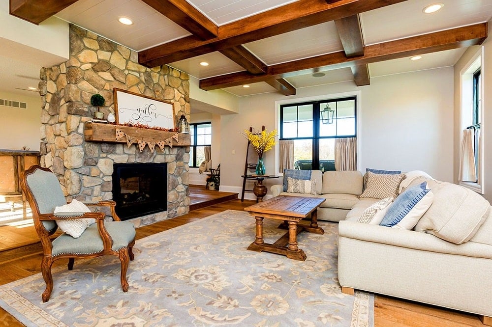 The living room has a large stone fireplace across from the large beige L-shaped sectional sofa topped with a beamed ceiling.