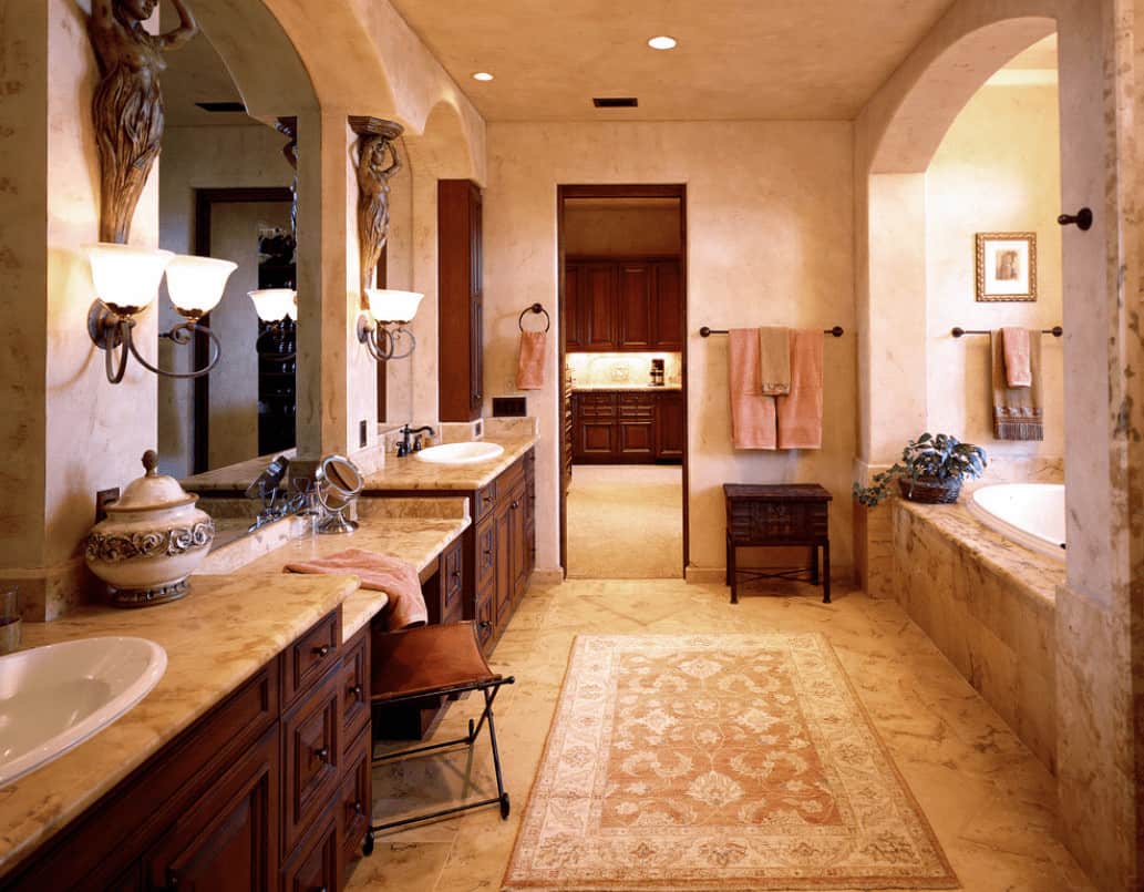A classic area rug lays on the tiled flooring in this beige primary bathroom showcasing alcove bathtub and dual sink vanity with marble countertops and frameless mirrors.