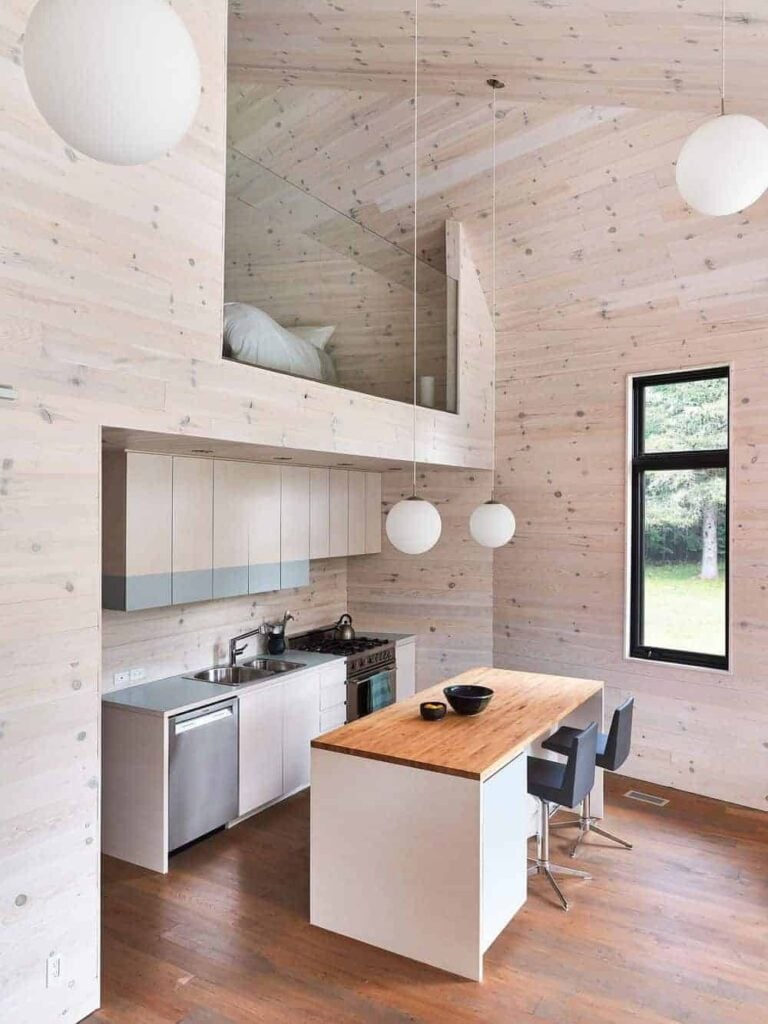 A breakfast island with black leather stools faces the light wood cabinets in this small kitchen with a glazed window and high wood plank ceiling mounted with globe pendant lights.