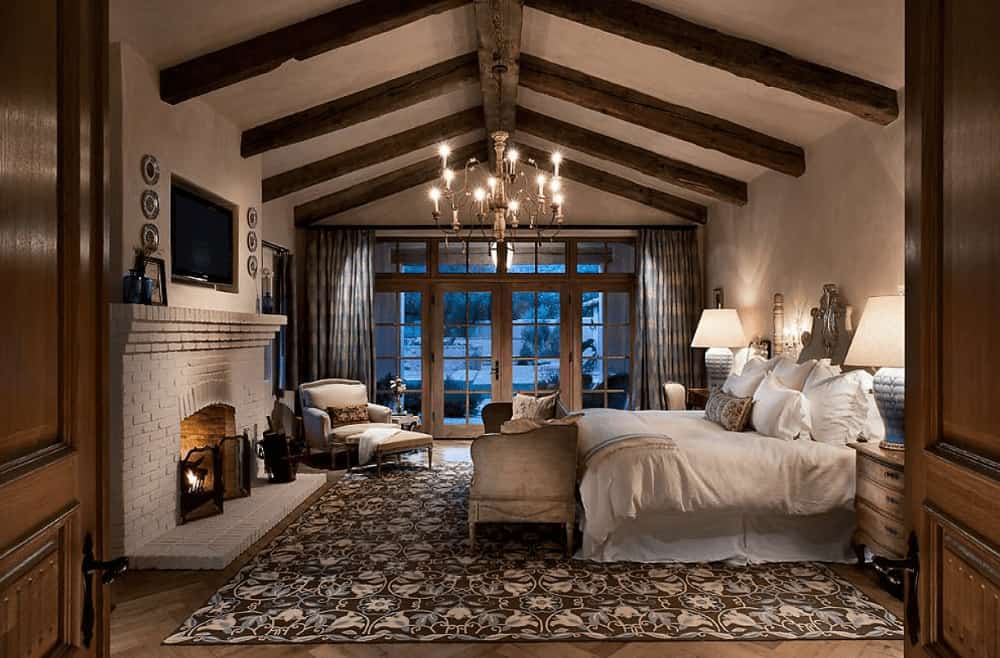 Cozy bedroom with a skirted bed on a floral rug and a brick fireplace with a television on top. It has herringbone flooring and wood beam ceiling mounted with a candle chandelier.