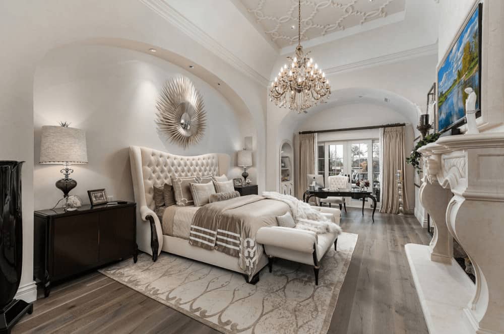 Mediterranean bedroom decorated with a sunburst mirror and fabulous chandelier that hung from the ornate tray ceiling. It has a carved fireplace and a tufted wingback bed with dark wood nightstands on its sides placed on the arched inset wall.