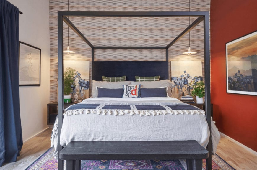 A purple floral rug lays on the light hardwood flooring in this bedroom with a canopy bed flanked by wooden nightstands and lovely artworks that are lighted by dome pendants.