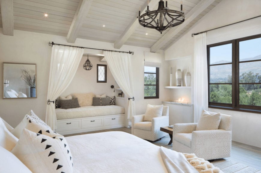 An all white bedroom with a comfy bed and armchairs along with a seat nook completed with white sheer curtains. It is illuminated by a vintage chandelier and recessed lights fitted on the wood beam ceiling.