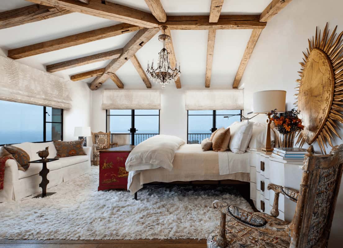 Mediterranean bedroom designed with a large sun decor and a fancy chandelier that hung from the wood beam ceiling. It has antique chairs and a skirted sofa facing the cozy bed that sits on a white fluffy rug.