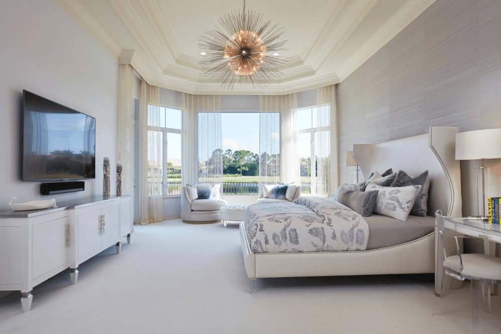 Sleek bedroom illuminated by a gorgeous sunburst chandelier that hung from the tray ceiling. It has round chairs and a wingback bed that faces the television mounted above the white stand.