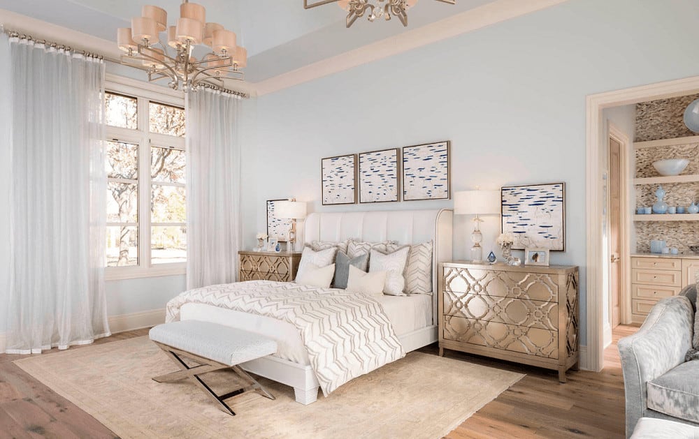 Light and airy bedroom with a white upholstered bed and classy nightstands topped with glass table lamps. It includes a lovely chandelier and sheer curtains covering the white framed windows.