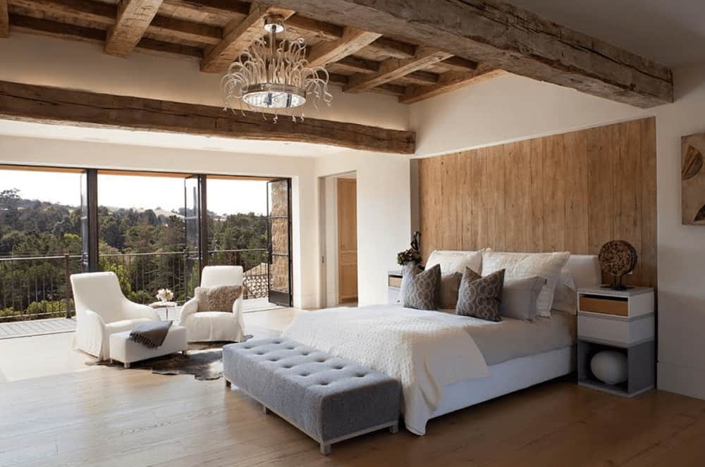 This bedroom showcases white seats and bed with a gray tufted bench on its end lighted by an eccentric chandelier that hung from the rustic ceiling.