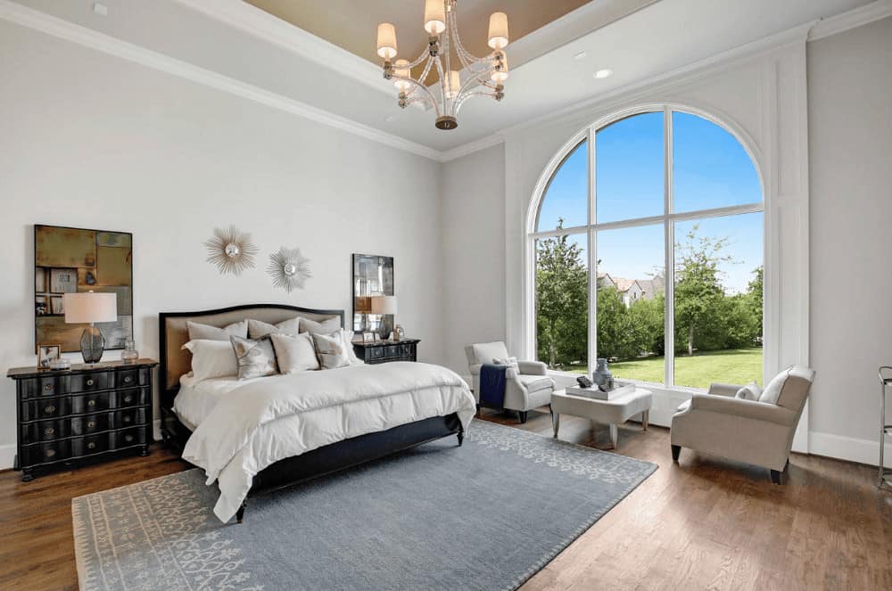 White bedroom decorated with a chandelier and sunburst mirrors mounted above the upholstered bed that's flanked by black nightstands and translucent table lamps. It has natural hardwood flooring and a large arched window framing the lush greenery.