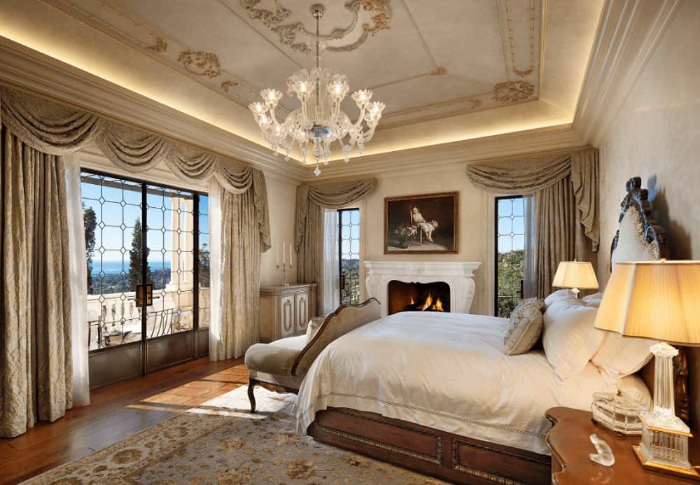 A fancy glass chandelier illuminates this deluxe bedroom offering a white fireplace and wooden bed that's surrounded with glazed windows dressed in classy drapes and valances.