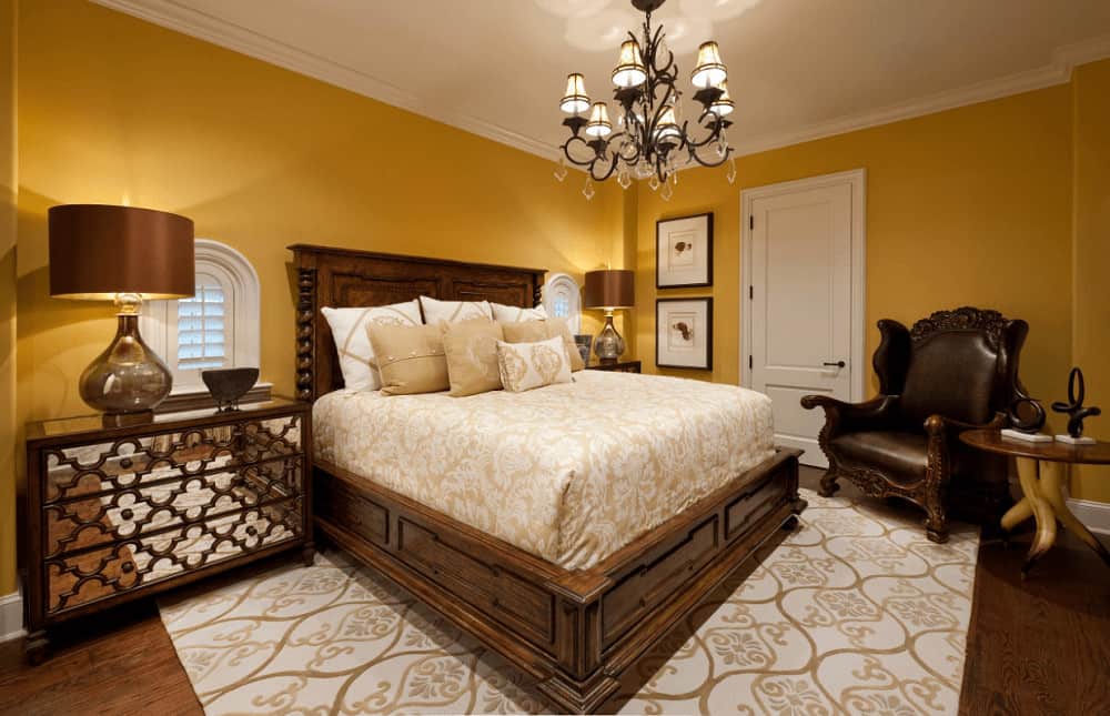 A black leather armchair faces the wooden bed on a patterned area rug over hardwood flooring. It is illuminated by  a wrought iron chandelier and glass table lamps that sit on mirrored nightstands.