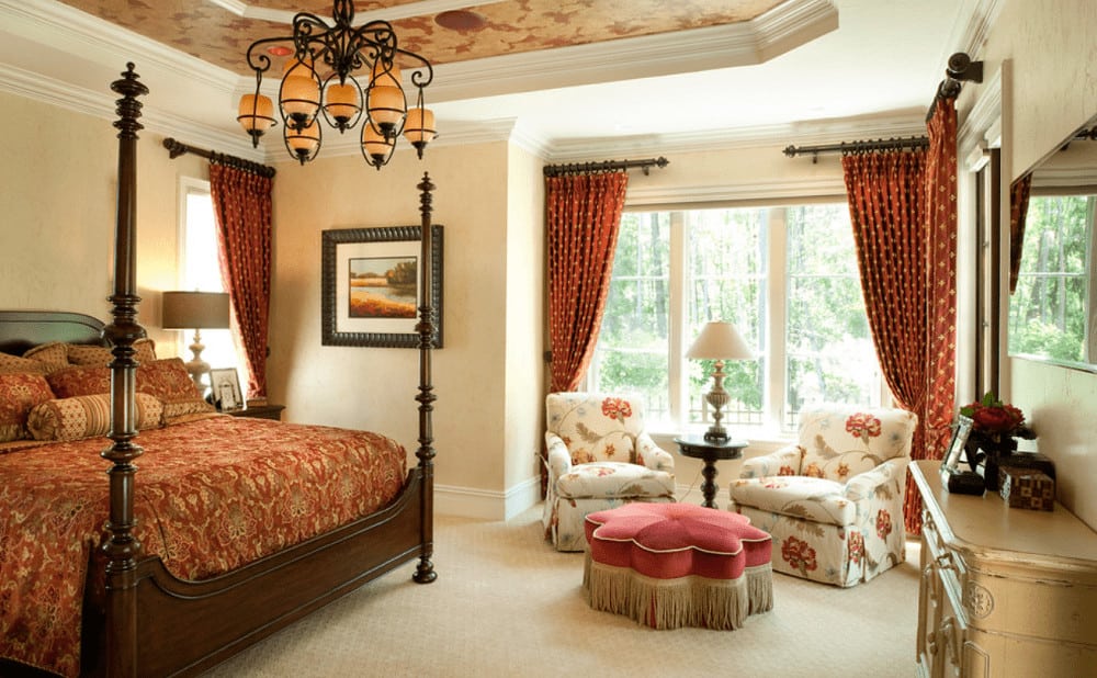 This bedroom showcases a seating area by the full height windows dressed in dotted red drapes along with a wooden four poster bed lighted by an ornate chandelier that hung from the tray ceiling.