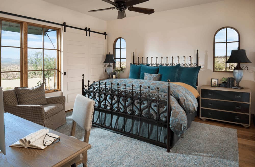 A gorgeous metal bed on a gray rug dressed in blue bedding stands out in this white bedroom with wide plank flooring and wooden framed windows that bring natural light in.