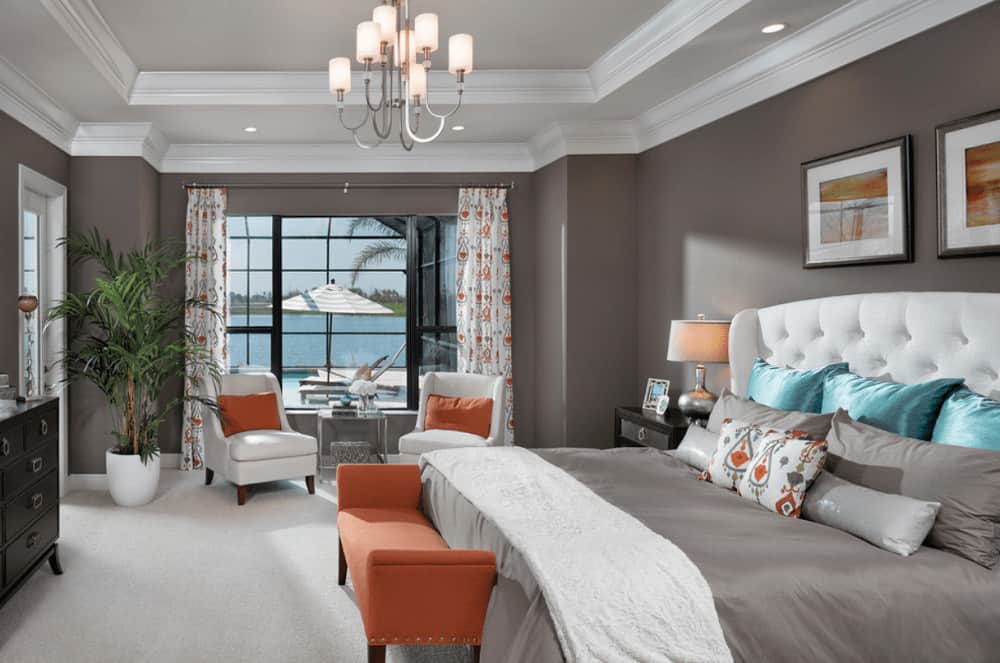 Orange pillows and bench add a warm accent in this gray bedroom featuring upholstered chairs and white tufted bed with black framed wall arts on top.