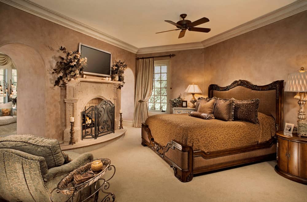 Warm bedroom with a carved wood bed and fireplace enclosed in a three-panel ornate fence. It includes a flat screen TV and an upholstered chair paired with a metal side table.