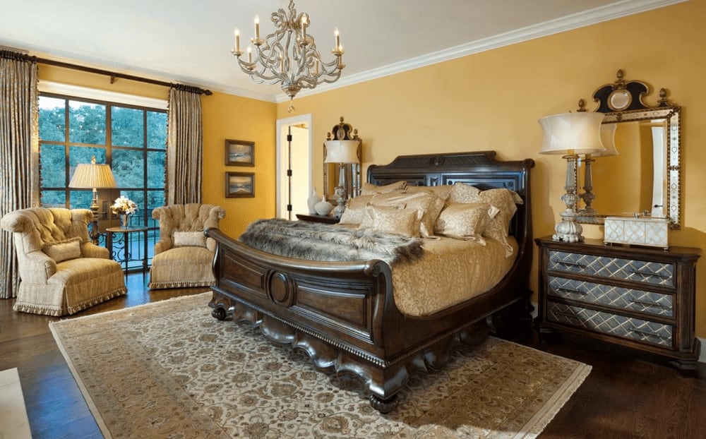 A stylish candle chandelier illuminates this yellow bedroom boasting tufted armchairs and a carved wood bed in between wooden nightstands paired with gorgeous mirrors.