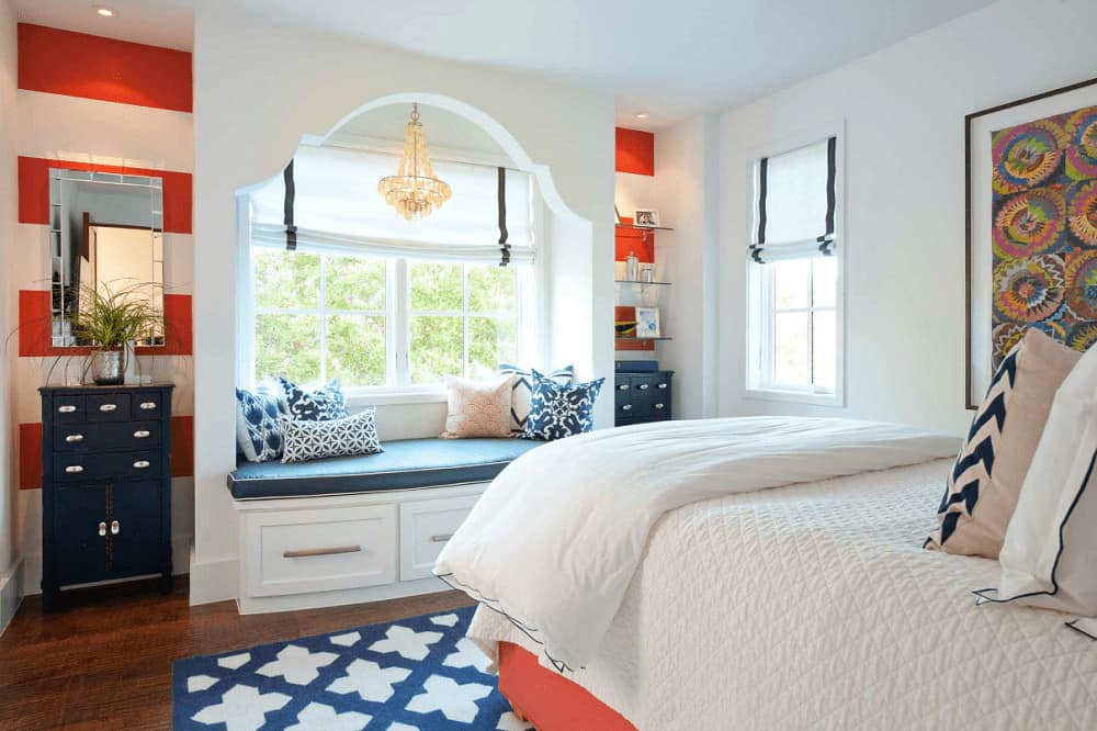 A comfy bed faces the alcove seat nook by the white framed windows lighted by a shell chandelier. It is placed in the middle of a red striped wall mounted with a mirror and floating glass shelves.