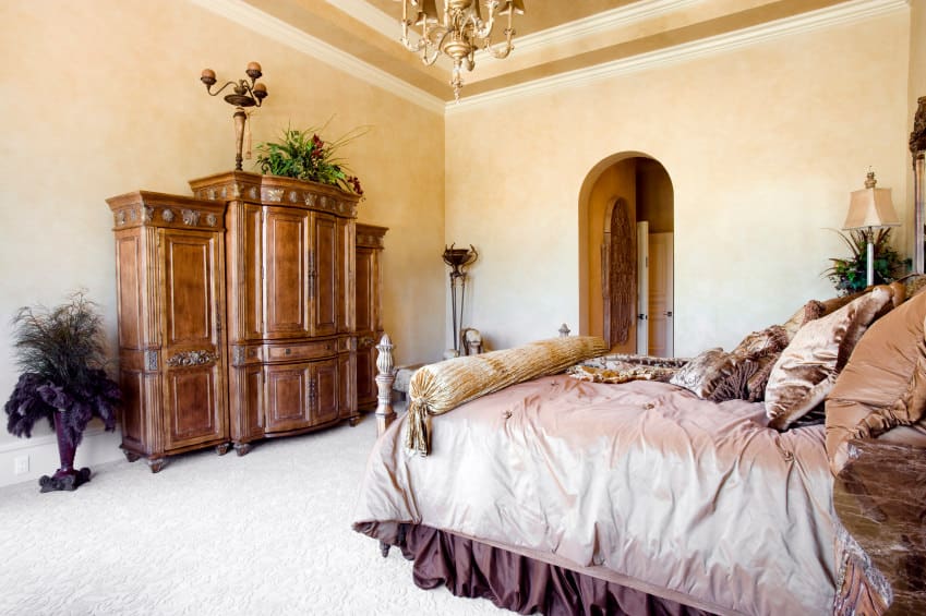 Mediterranean style primary bedroom with a wooden cabinet and a large cozy bed. The room is lighted by a gorgeous chandelier hanging from the tray ceiling.