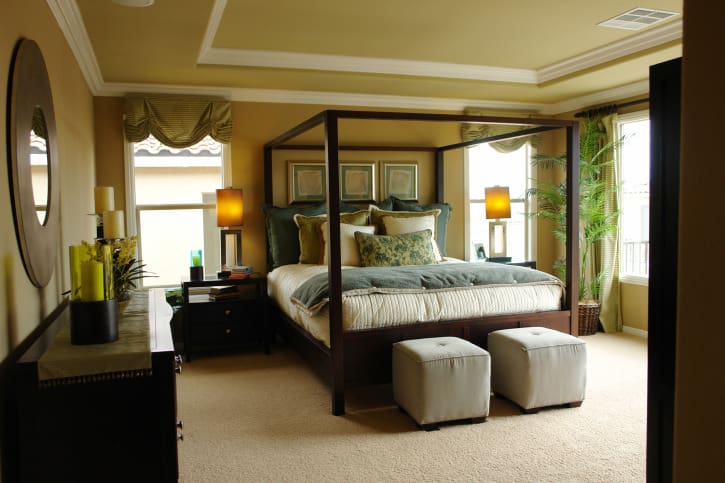 Country-style primary bedroom with yellow walls and a tray ceiling, along with carpeted flooring. The bed setup is lighted by table lamps on both sides.