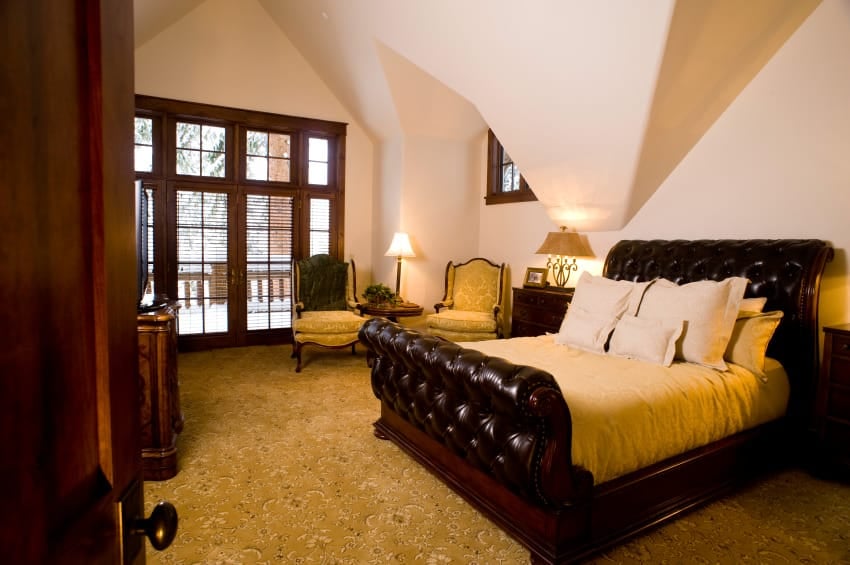 A medium-sized primary bedroom with a custom ceiling and gorgeous decorated carpet flooring. The room offers a luxurious bed, along with a sitting area on the side.