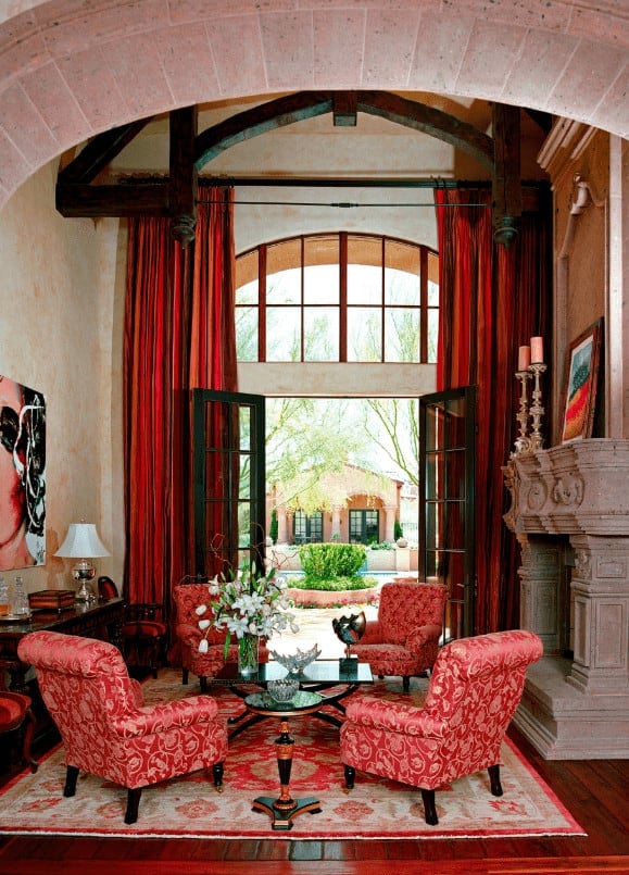 This living room boasts a concrete fireplace and black coffee table surrounded by red floral armchairs. It has wide plank flooring and a French door that opens to the yard.