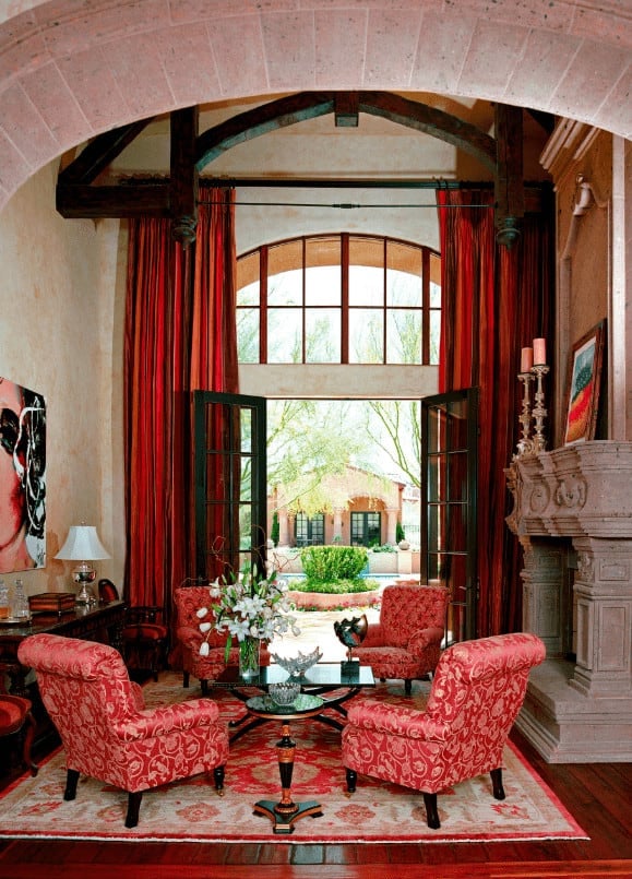 Floors & Walls: Here we have another extravagant room that is taller than it is wide, which is a refreshing sight when so many rooms are dedicated to being horizontally spacious. Gorgeous cherry wood floors are complimented by a stone fireplace, and incredible stucco walls that guide you towards an impressive vaulted ceiling complete with exposed beams and a stone  archway.  Furniture: Red is way more present in the furniture of this room but still holds its dominance. Patterned red accent chairs, a patterned red area rug, luxurious red curtains, and red themed art bring the room together without being too overbearing. Decor: With such bold pieces of furniture, it feels safer to use minimal decorations which we see here in the form of a huge vase of white flowers, crystal bowls and candle holders, and a simple lamp. Where a lot of the decoration comes from is the beautiful view of the courtyard just beyond the french doors. This stunning living space was designed by Robert Burg Design.