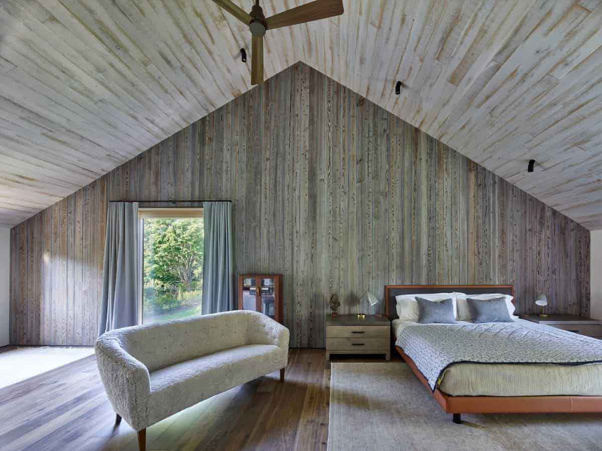 This large Rustic-style bedroom has a wooden cathedral ceiling that blends with the wooden walls and hardwood flooring. This envelops the bed and the cushioned couch that matches with the area rug into a charming embrace of wooden elements.