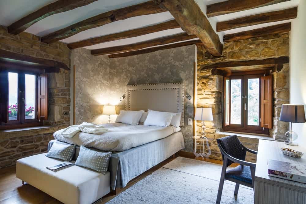 The white ceiling is adorned with beautiful irregular textured wooden beams that set a unique aesthetic to this Rustic-style bedroom that has rough stone walls and hardwood flooring that gives emphasis on the beige platform bed.