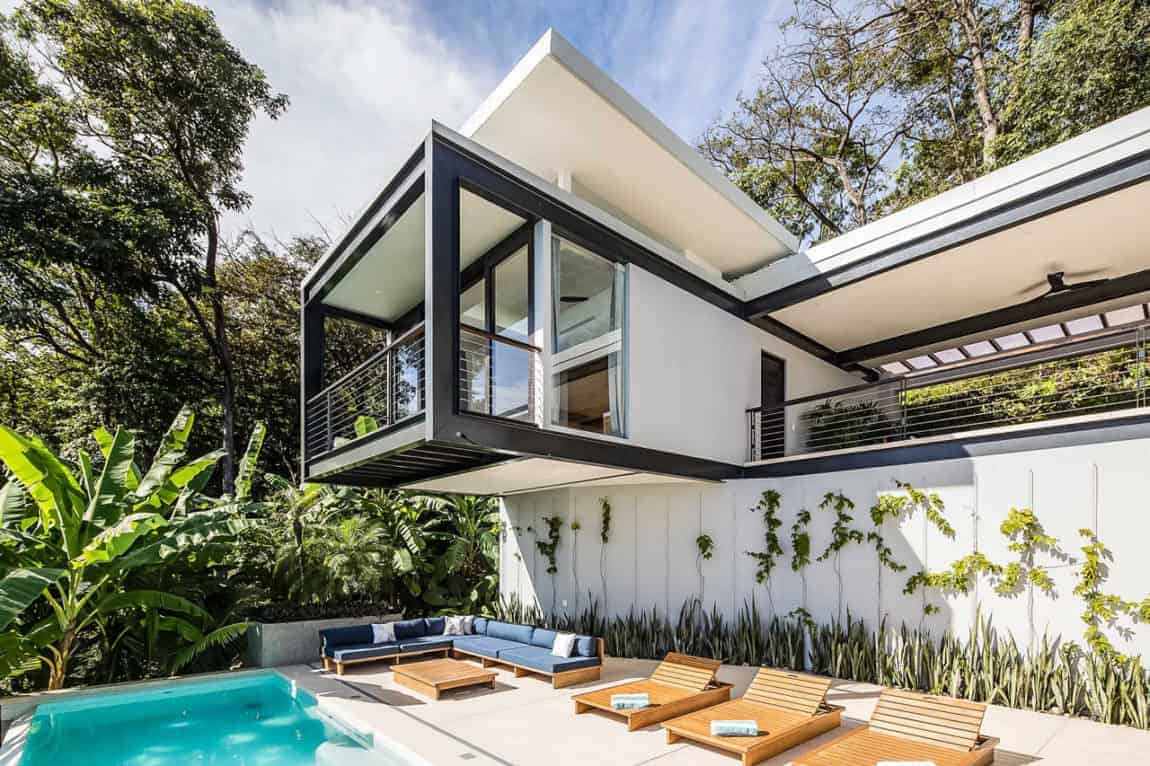 This is a beautiful pool side scene accented with a distinct Tropical-style landscaping. The swimming pool that stands out against the bleached concrete flooring that has an L-shaped wooden sofa along with three wooden lawn chairs. This has a background of white walls with creeping plants on it.