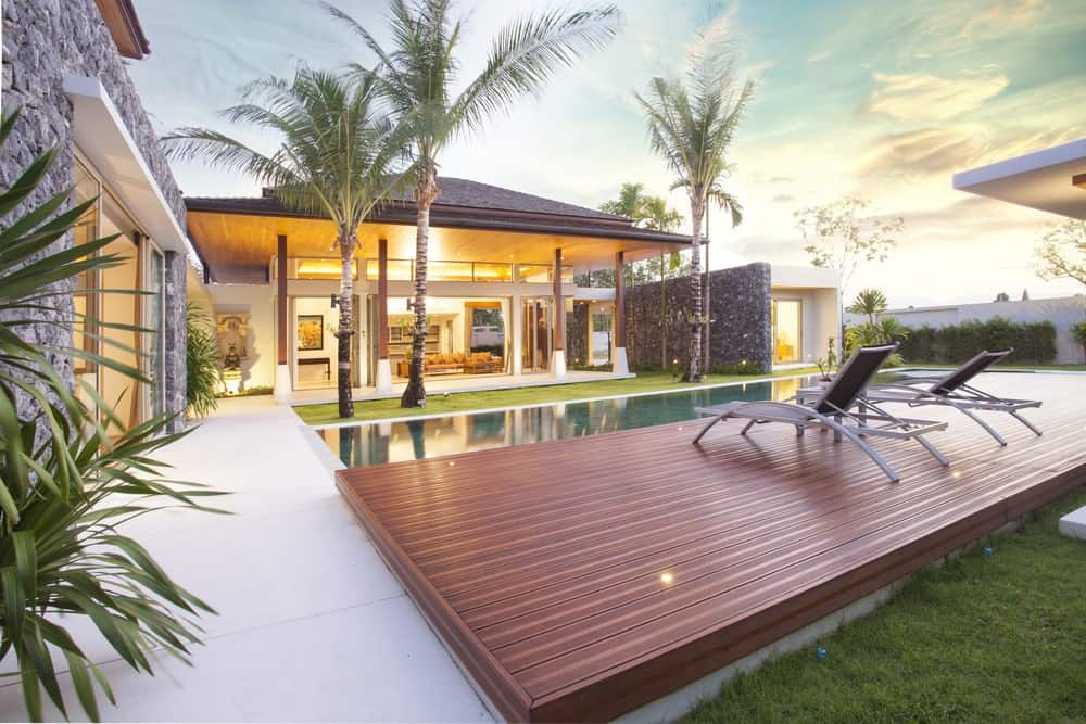 This relaxing Tropical-style landscape has a rectangular pool that is flanked one side by a wooden platform with lawn chairs on it. The other side of this pool has a small carpet of grass with three tall tropical trees that adorns the lovely home.