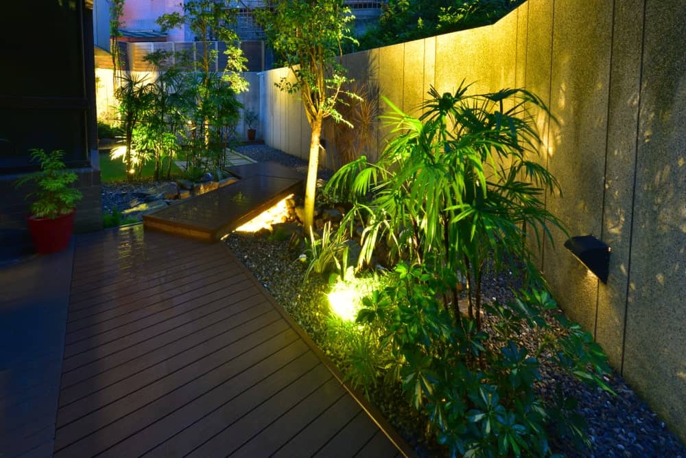 This is the view of the side of the house that has a beautiful wooden walkway adorned with a distinct Tropical-style landscape that has various tropical plants that is lit with warm yellow lights to emphasize the shadows and silhouettes of the leaves on the surrounding concrete fence.