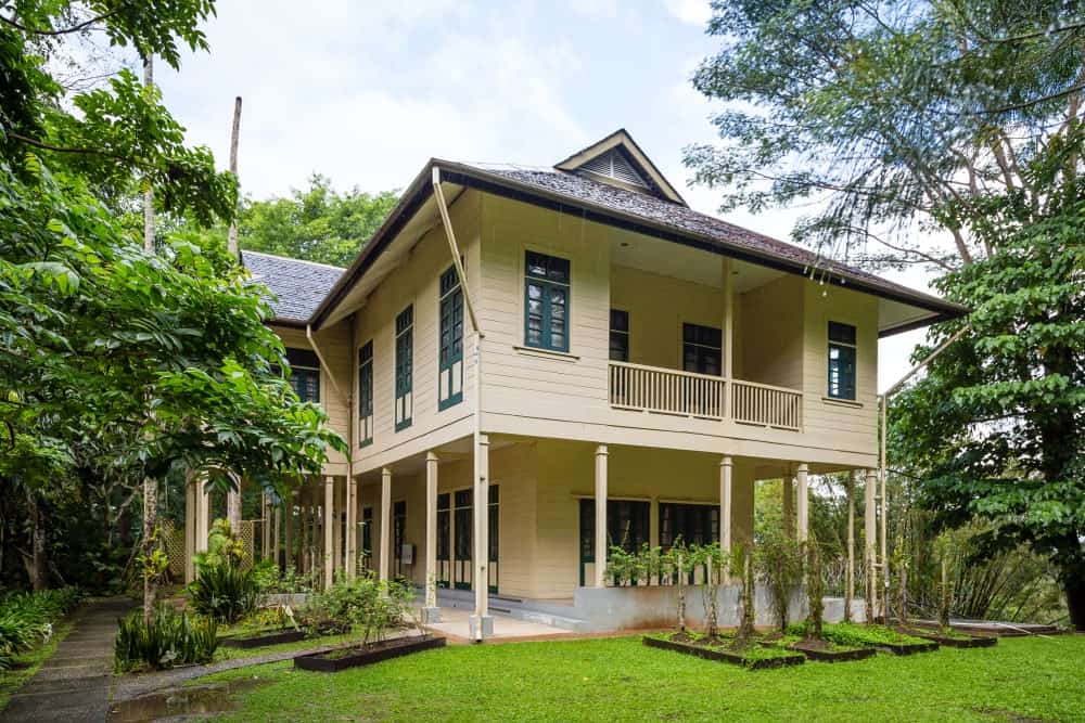 This is a charming wooden home given a Tropical-style landscape to complement its beige exterior walls. There are various tropical trees and shrubberies with some taller than the house and some accenting the concrete walkways on the side.