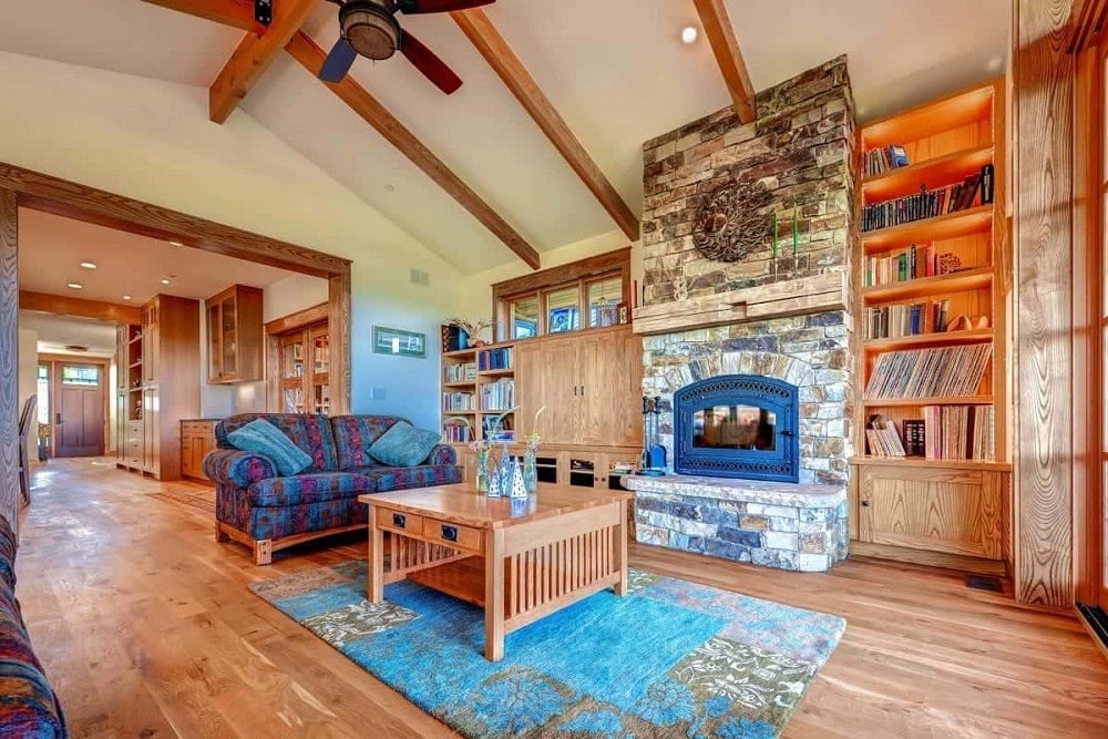 This is a view of the living room with a tall stone structure that houses the fireplace and supports the built-in wooden cabinets and shelves on the sides across from the colorful sofa and patterned area rug that makes the simple wooden coffee table stand out.