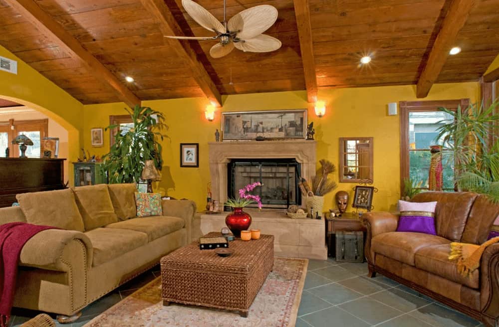 A brass ceiling fan hangs over the wicker ottoman surrounded by cozy seats and a concrete fireplace topped with a framed wall art.