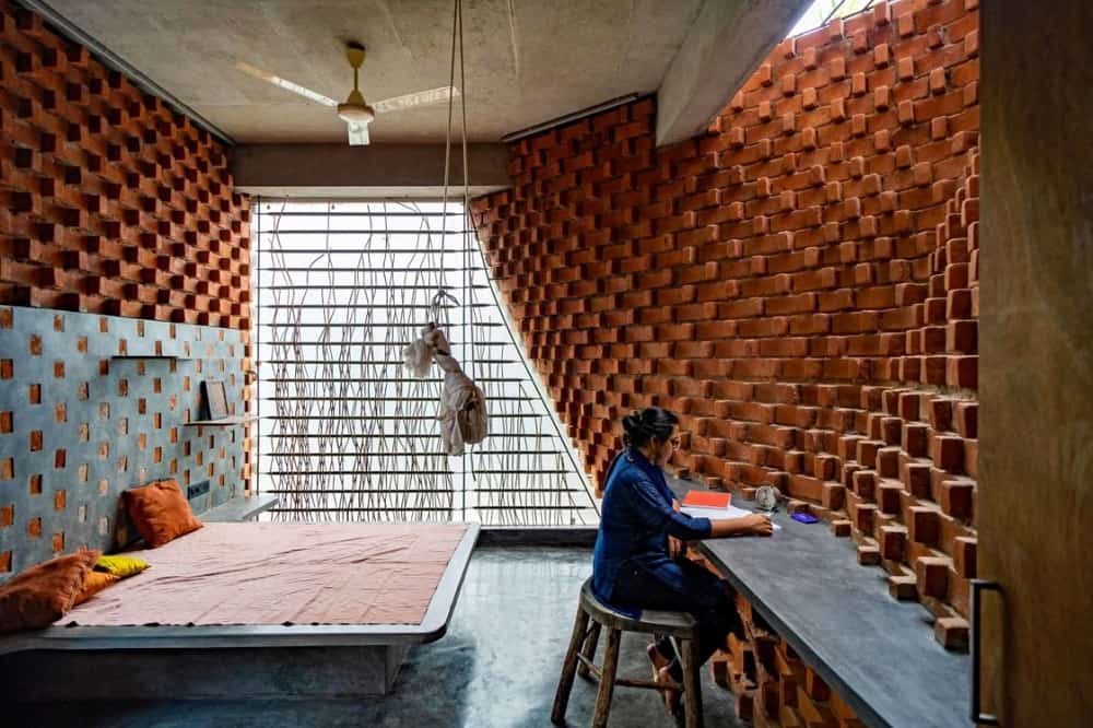 This is a full view of the bedroom that has a curved and textured red brick wall with a built-in concrete desk on the other side of the concrete platform bed.